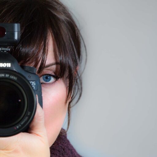 Close-up self-portrait of photographer Fiona Whittaker holding a DSLR camera, with one blue eye visible beside the lens against a plain background.