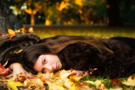 Carlotta lying beneath a tree surrounded by fallen autumn leaves in warm golden light. Portrait of a young woman named Carlotta lying on autumn leaves beneath a tree, wearing a dark fur coat in warm golden evening light.