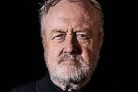 A close-up portrait of Graham Whittaker captured in the final years of his life. Close-up portrait of Graham Whittaker with a grey beard and blue eyes, photographed against a dark background with dramatic lighting.