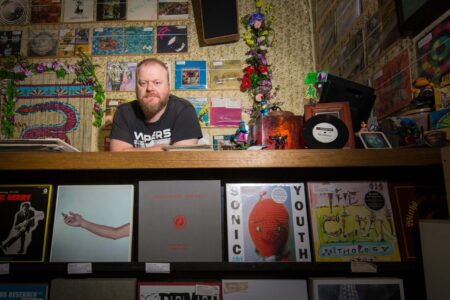 A record store owner standing behind the counter of his vinyl shop, surrounded by album covers and music memorabilia. Portrait of a bearded man behind the counter of an independent record store surrounded by vinyl records, album covers and music memorabilia.