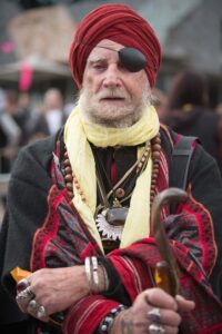 “The Wizard” photographed in richly layered clothing and ceremonial accessories during a public gathering. Portrait of a man known as “The Wizard” wearing a red turban, eye patch, patterned robes and jewellery while holding a walking cane in a crowded outdoor setting.